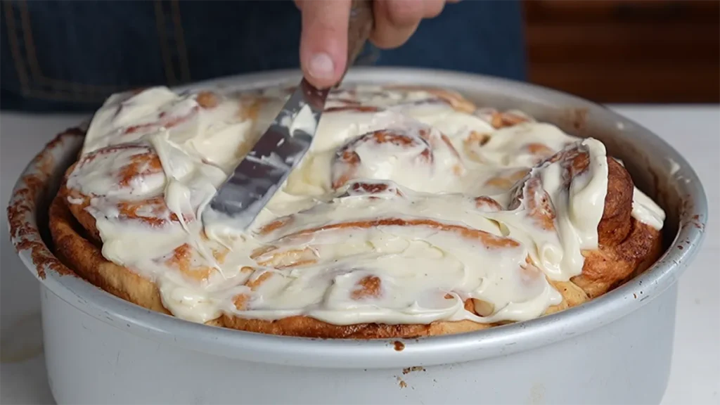 Spreading cream cheese frosting on baked cinnamon rolls.