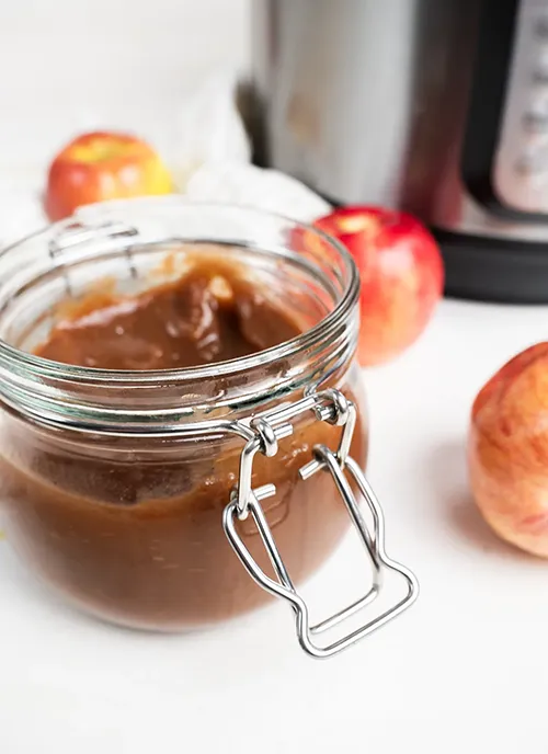 Jar of apple butter, with a few apples and an Instant Pot pressure cooker in the background.