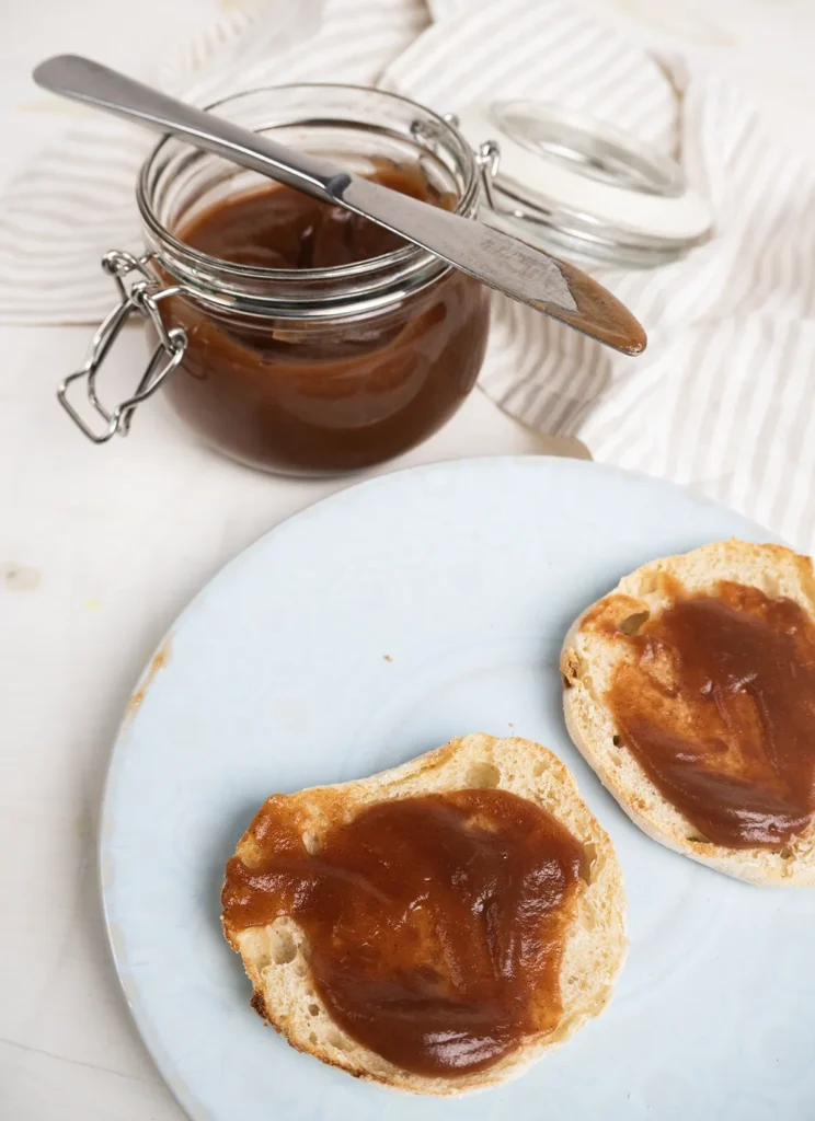Apple butter spread on toasted english muffins on a plate. An open jar of apple butter is in the background.