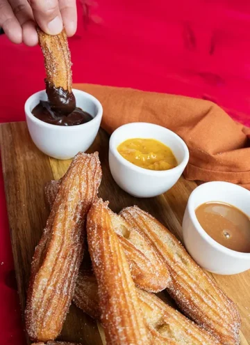 A pile of sourdough churros on a wooden board. In the background, a hand is dipping a churro into some spiced chocolate dip.