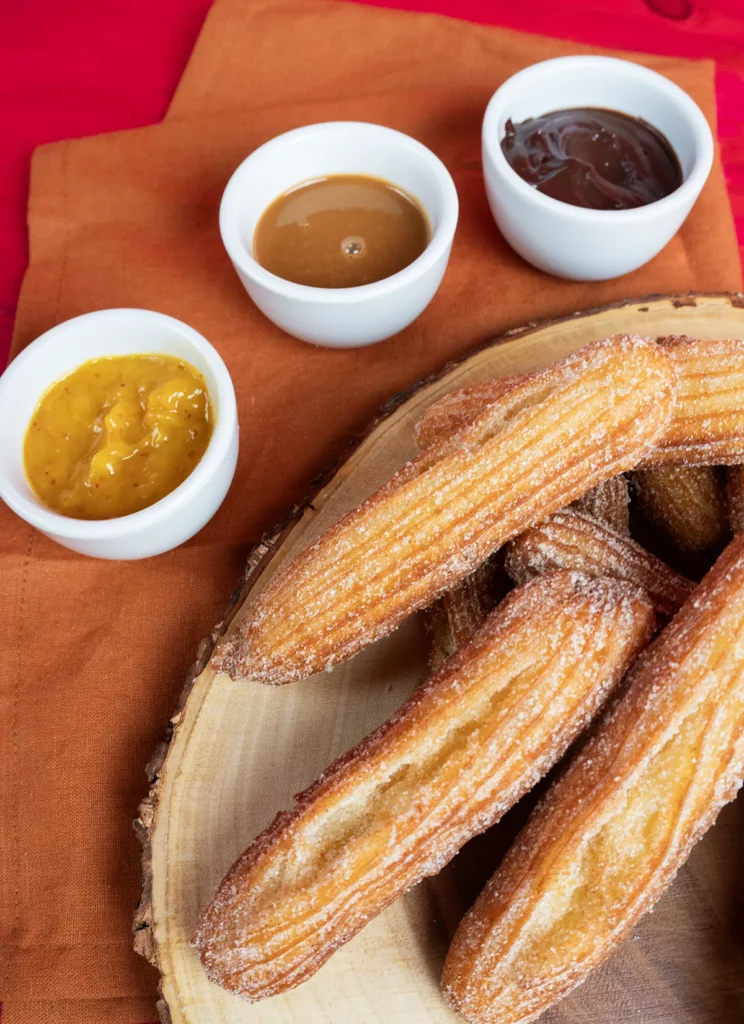 A stack of sourdough churros on a wooden platter alongside three dipping sauces.