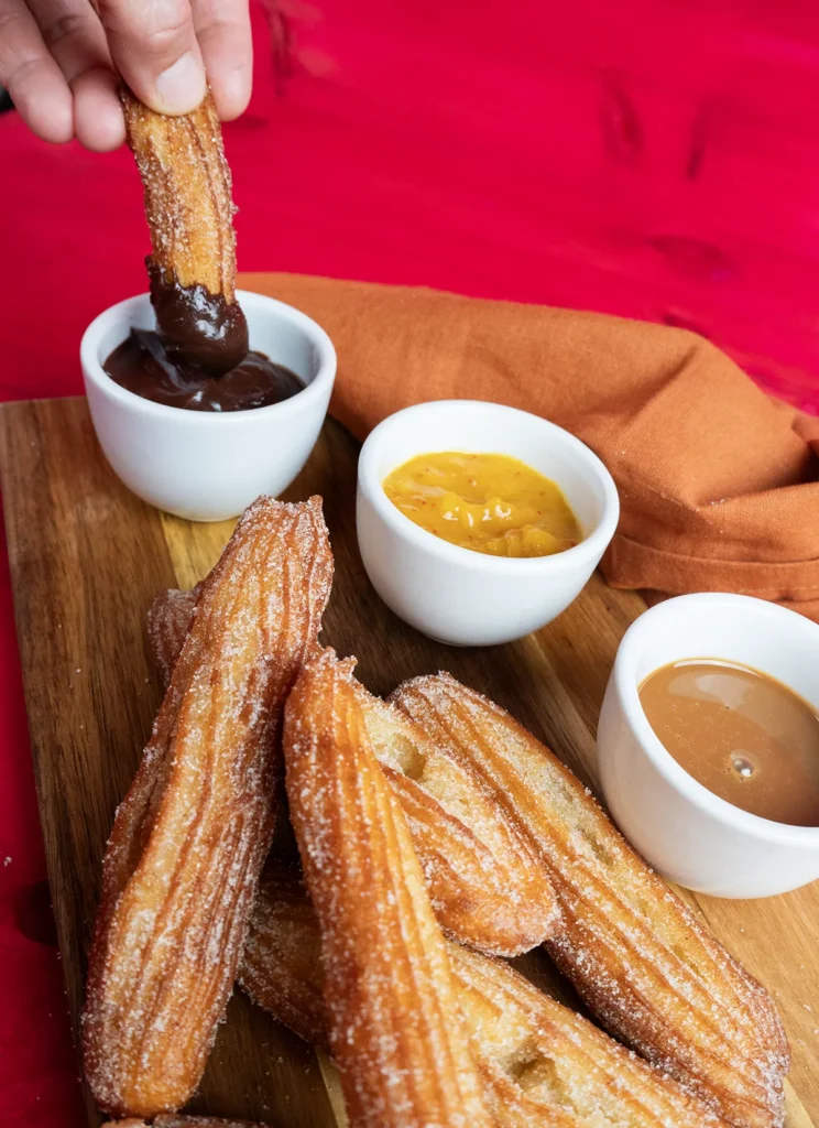 Dipping a sourdough churro into some spiced chocolate ganache. On a wooden platter with a stack of sourdough churros.