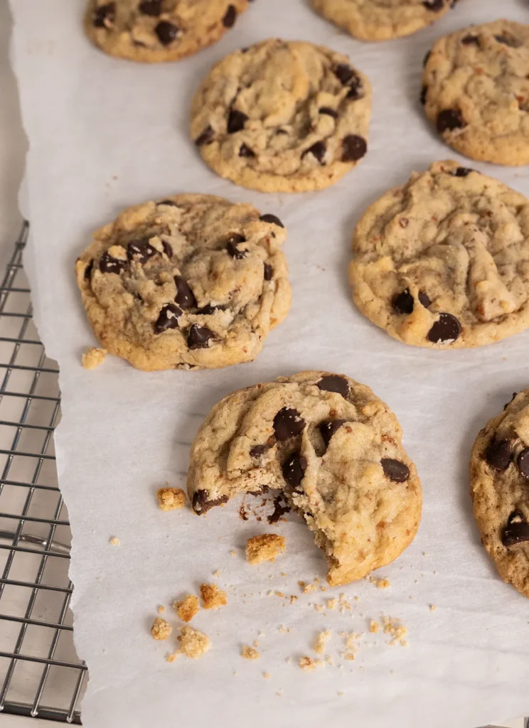 Top view of sourdough chocolate chip cookies laid out on parchment paper with crumbs. One cookie has a bite taken out of it.
