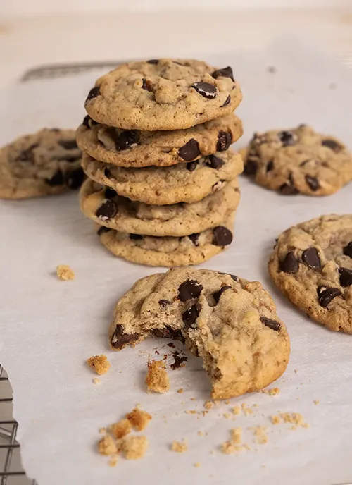 Stack of sourdough chocolate chip cookies on parchment paper with crumbs. One cookie in the foreground has a bite taken out of it.