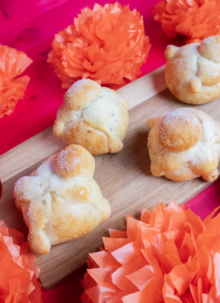 Sourdough Pan de muerto rolls lined up on a wooden board, with orange flowers in the background.
