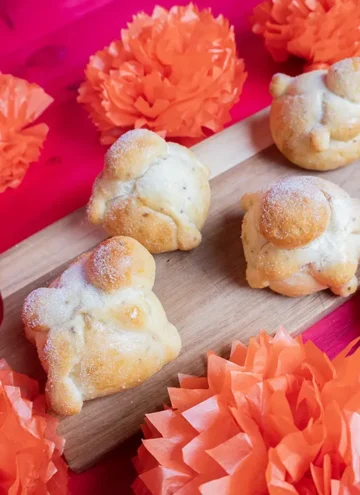 Wooden board with sourdough Pan de Muerto lined up, and orange flowers in the background.