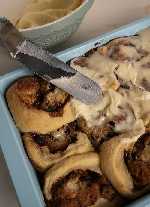 A pan of cookie dough cinnamon rolls being slathered with cream cheese frosting.