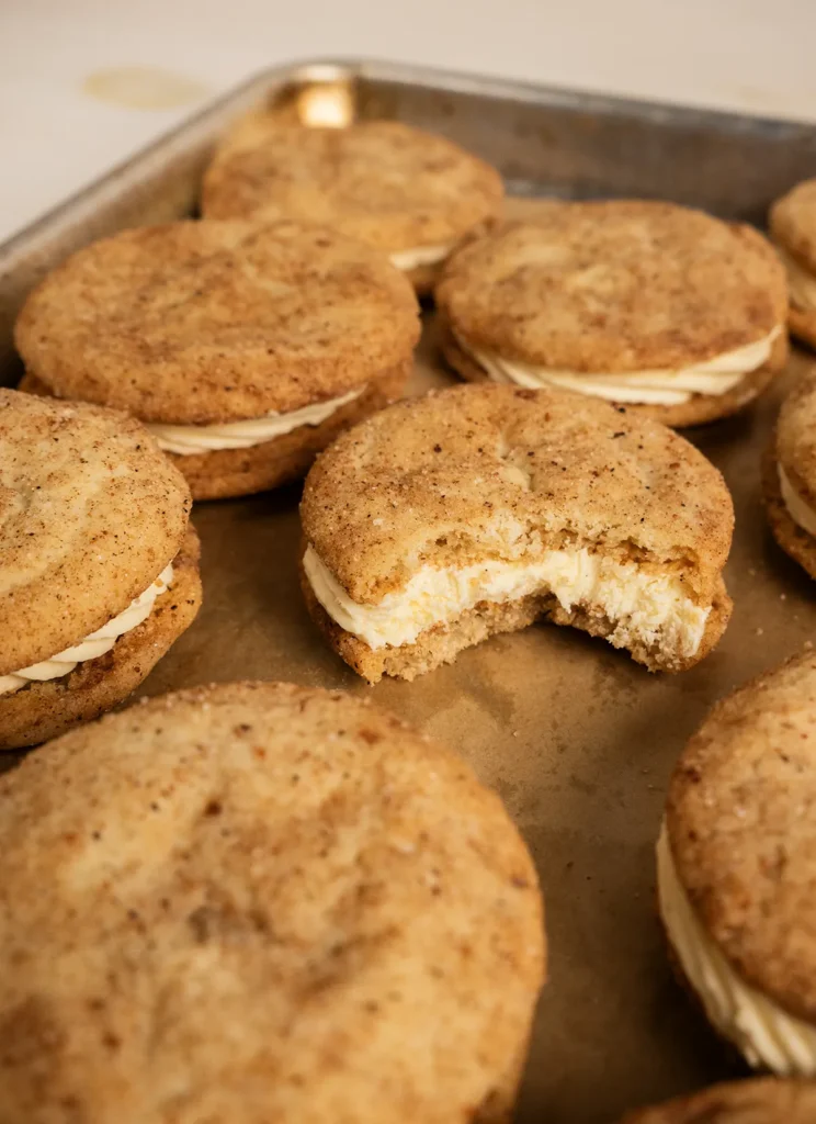 Eggnog snickerdoodle sandwich cookies on a baking sheet. One has a bite taken out of it.